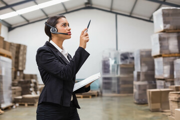 Warehouse supervisor with headset mic inspecting stock using clipboard pen amid pallet box stacks