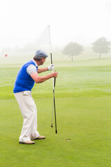 Senior golfer standing on manicured putting green, lifting flagstick from hole in misty morning