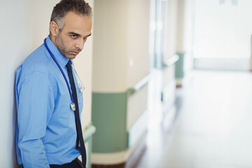 Middle Eastern male doctor leaning against wall in hospital corridor with stethoscope, copy space