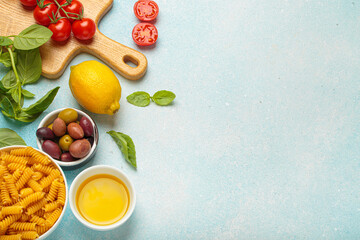 Cherry tomatoes, basil, olives, lemon, olive oil, fusilli pasta and wooden cutting board are creating a frame with copy space on a light blue background, concept of Italian cuisine cooking, copy space
