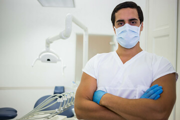 Male dentist standing wearing surgical mask at clinic with dental lamp and gloves, copy space