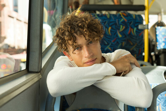 Early twenties male passenger leaning on bus folding arms over patterned seat gazing through window