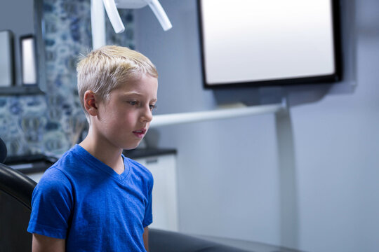 Male child sitting in dental treatment chair wearing blue T-shirt under overhead lamp with monitor