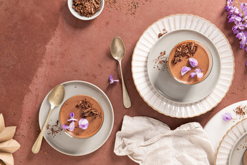 An overhead view shows two creamy chocolate mousses in glasses, styled with spoons, garnishes, and decorations on a textured surface, inviting treat.