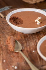 Individual servings of smooth chocolate mousse in white bowls, one with a spoonful being lifted, garnished with tiny white flowers, set on a rustic wooden board