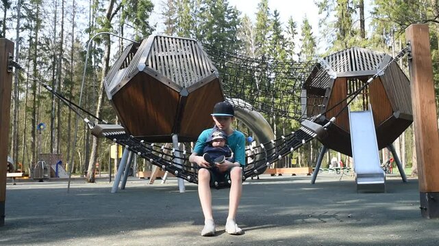 A 12 year old boy with his baby brother on his knees swings on a hammock swing at a playground in Borovsky Park in Moscow.	