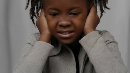 Distressed young girl covering her ears with hands, showing discomfort or pain with emotional expression, wearing neutral colored sweater in a studio setting - Powered by Adobe