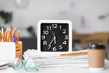 Alarm clock with papers, holder with pencils and cup of coffee on table in office