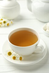 Delicious chamomile tea in cup, fresh flowers and teapot on white wooden table, closeup