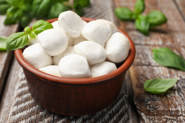 Tasty mozzarella cheese balls in bowl and basil on wooden table, closeup
