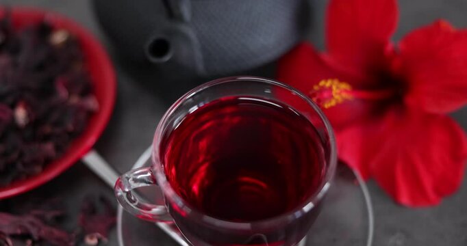 Tasty hibiscus tea in cup, teapot, dry roselle sepals and beautiful flower on grey table, camera moving