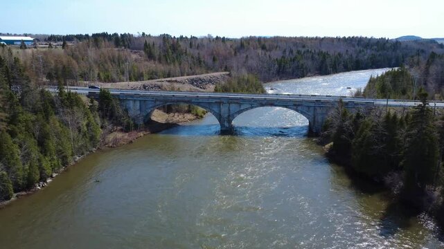 Drone footage flying above the swollen Mitis River during spring freshet and concluding with a pass under the Arthur‑Bergeron concrete arch, Quebec, Canada, 2025.