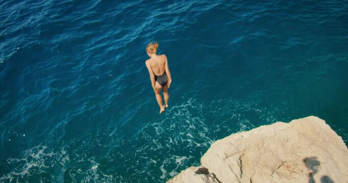 Young woman in swimsuit jumps off rocky cliff into deep blue sea on sunny day