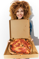 Happy young woman with curly hair holding a large pepperoni pizza in a takeout box against a bright...