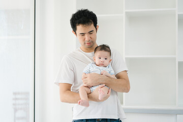 Fototapeta premium Asian devoted young father lovingly holds his three-month-old baby in a warm, inviting room. Their close bond radiates affection, peaceful atmosphere of comfort, safety, and unconditional love.