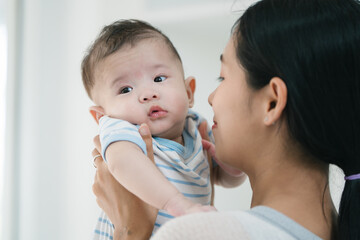 Asian loving young mother gently embraces her three-month-old baby in a cozy room, radiating warmth and affection. Their tender connection fills the space with a sense of comfort, security, and love.