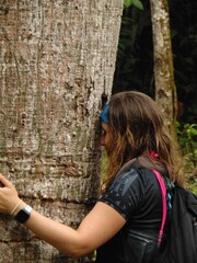 A woman with dark skin, light hair, and a black blouse is hugging a large tree while showing respect and affection towards it.