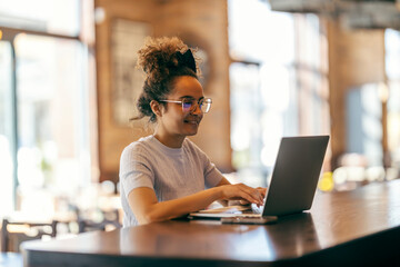 Cheerful multicultural female freelancer sitting in coffee shop and typing project on a laptop...