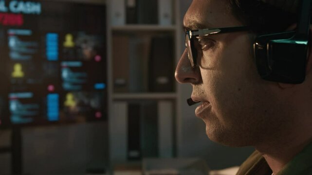 Side close-up of face of Asian male fraudster wearing headset with microphone threatening victim while working from scam call center, blackmailing into transferring funds into criminals account