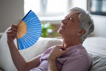 Lack of cooling in summer, heatstroke risk for elderly people. Senior man using fan to survive sweltering conditions, extreme weather vulnerability, suffer from unbearable temperatures at home