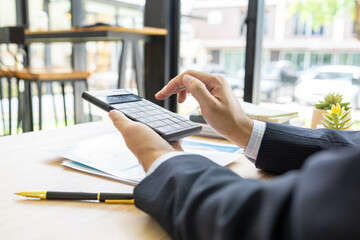 A man in a suit is sitting at a table with a piece of paper in front of him. He is looking at the paper and calculating and comparing data on the document, his phone, and a calculator.


