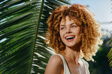 smiling woman with curly hair enjoying sunny tropical outdoors, natural light, green palm leaves,...