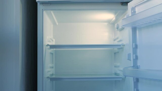 An empty refrigerator with no food, a hand opens and closes the home fridge with empty shelves meant for groceries
