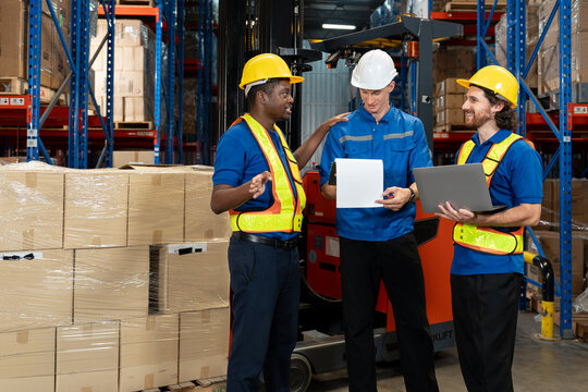 Three male warehouse employees, different ethnicities, in uniform and helmet, discussing work details using document and laptop while standing beside forklift and stacks of cardboard boxes.