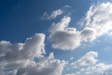 Soft, fluffy clouds float lazily in a clear blue sky during the day. Sunlight illuminates the white clouds, creating a serene visual experience perfect for daydreaming or inspiration.