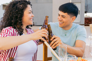Young Latin man and woman gaze lovingly at each other and toast with beer bottles on a terrace date