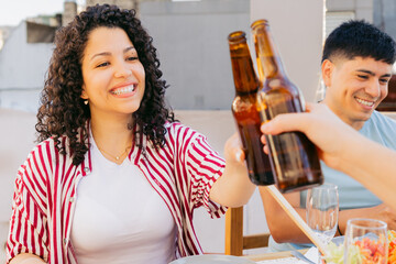 Young Latin woman, sitting with friends, toasting with beer bottles and smiling on terrace at sunset