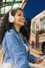 Independent young latin woman, smiling, listening music with headphones, waiting for the bus alone at night