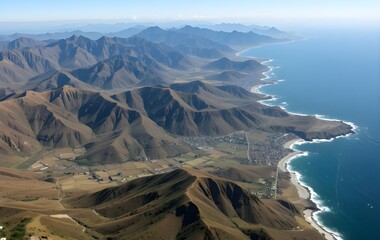 Aerial view of Baja California’s dramatic coastline, showcasing steep mountains, coastal towns, and curving beaches meeting the Pacific Ocean under a clear sky.