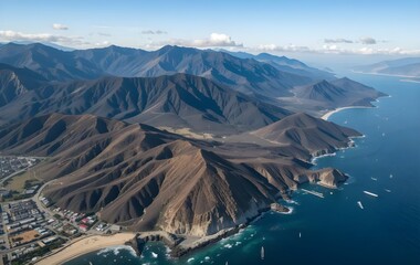 Aerial view of coastal mountains meeting the Pacific Ocean in Baja California, Mexico. Rugged terrain, deep blue sea, and scattered boats create a dramatic and scenic landscape.