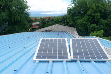 Two large solar panels are installed on a blue metal roof of a rural house using steel legs to support them.
