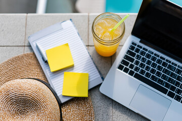 Overhead view of summer remote working scene: laptop, e-reader, sticky notes, iced drink, straw hat on tiled balcony floor, 