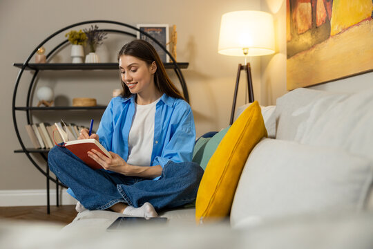 Young woman sitting comfortably on living room couch while writing in notebook - Powered by Adobe