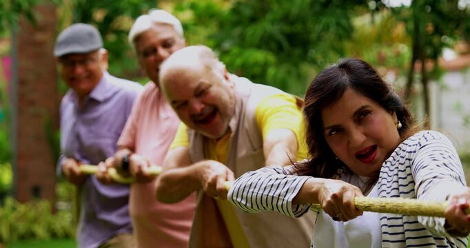 Senior playing tug of war in garden, Indian Asian elderly friends enjoying outdoor physical activity with rope, laughing and pulling together as part of fun reunion game amid green nature