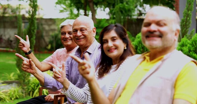Indian seniors pointing outdoors, Asian elderly friends seated in a row against lush green garden background, smiling and showing thumbs up with positive expressions looking at camera