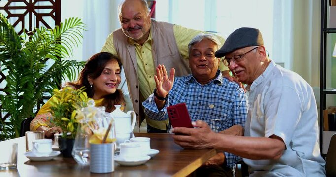 Seniors video calling at dining table, Indian Asian old age friends reuniting at home, attending group call using smartphone while enjoying tea, snacks, friendly talk at modern indoor dining setup