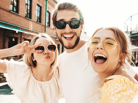 Group of young three stylish friends in the street.Man and two cute female dressed in casual summer clothes.Smiling models having fun in sunglasses.Women and guy making photo selfie on smartphone