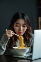 Young woman eating instant noodles while working at laptop indoors