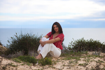 Beautiful young brunette woman is barefoot and sitting on the dunes by the beach. The woman is...