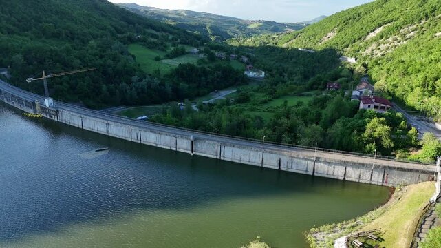 Aerial view of Mignano Dam, capturing its sweeping concrete arc, tranquil reservoir, and the lush, rolling hillsides of the Arda valley, dotted with houses and winding rural roads, PC, Italy
