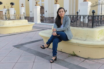 A woman with long dark hair, wearing a checkered blazer and jeans, sits calmly on a pale yellow bench © sandor