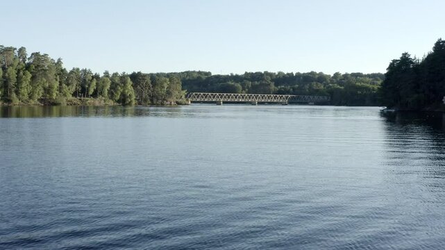 Aerial view flying over calm lake towards Pont de Lantourne bridge in distance, forested shores, Marcillac La Croisille, France