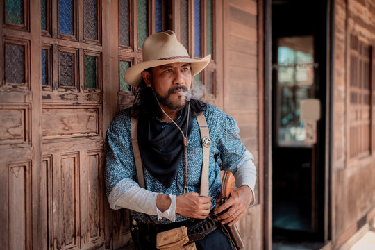 Portrait elder cowboy standing at a wooden bar, aiming revolver upward in a dramatic on the weapon suggest respond to danger, possibly during a shootout or high-stakes standoff.