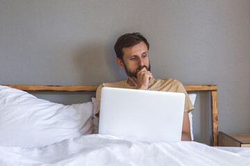 Man sits in bed with laptop, looking thoughtful and focused, working in a calm home setting.