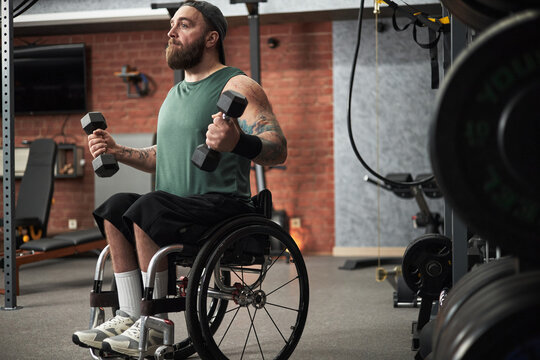 Man in wheelchair lifting dumbbells in gym setting, demonstrating strength and determination. Background includes gym equipment and weights