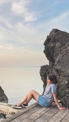 A young woman relaxes on a wooden deck by the sea, surrounded by rocky cliffs and a calm pastel sky during sunset. vertical1
